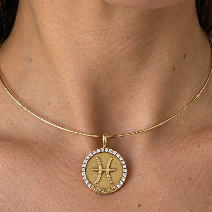 A close-up of a model's neck and chest wearing a delicate gold bangle necklace with a circular Pisces zodiac sign pendant adorned with small clear stones.