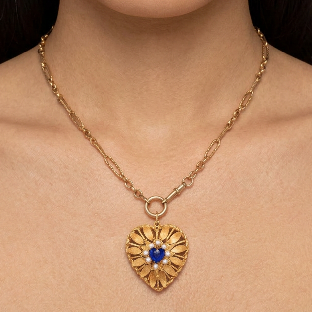 A close-up of a model wearing a gold heart-shaped pendant necklace with a central blue gemstone, set against a neutral gray background.
